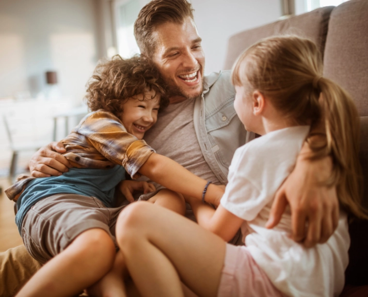 Smiling father sitting on the floor laughing and hugging his two children, showing love and family connection