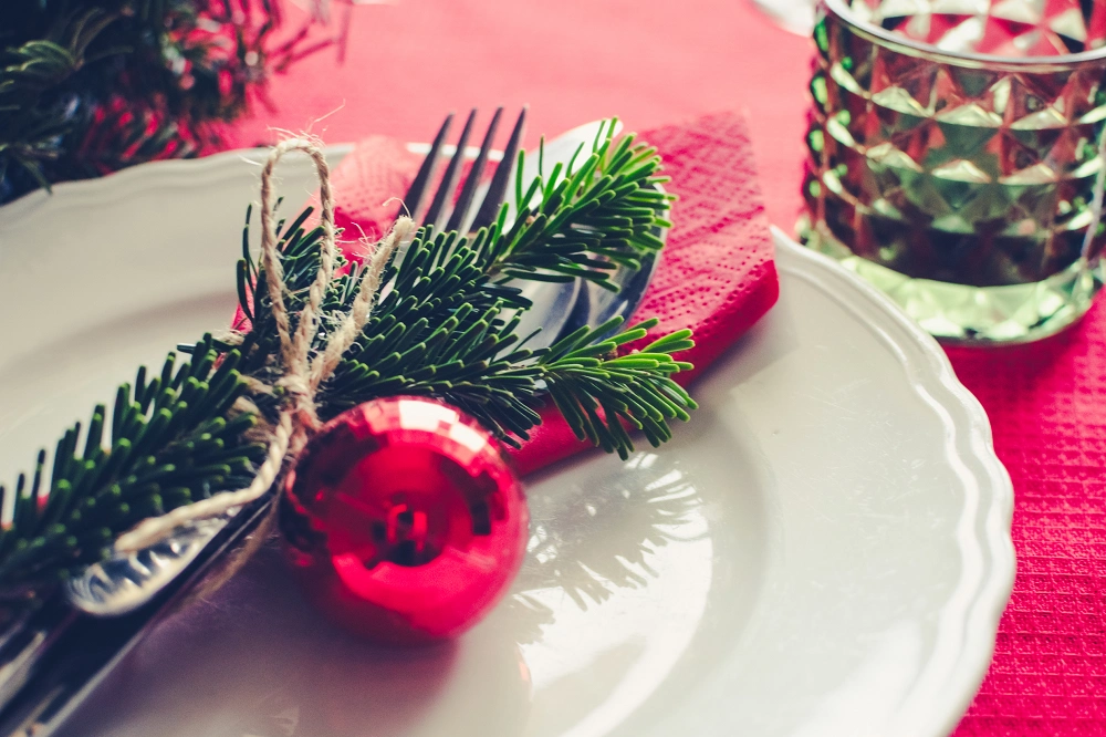 Christmas table setting with fork and spoon wrapped in pine and twine beside a red ornament on a white plate