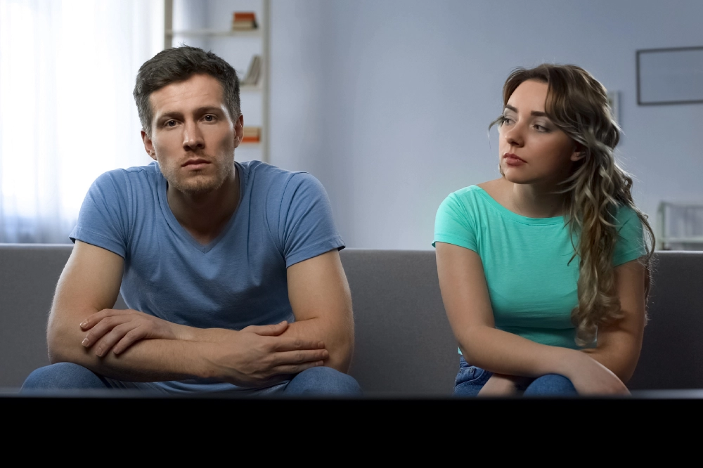 Couple sitting on couch with serious expressions, suggesting tension and emotional distance in relationship