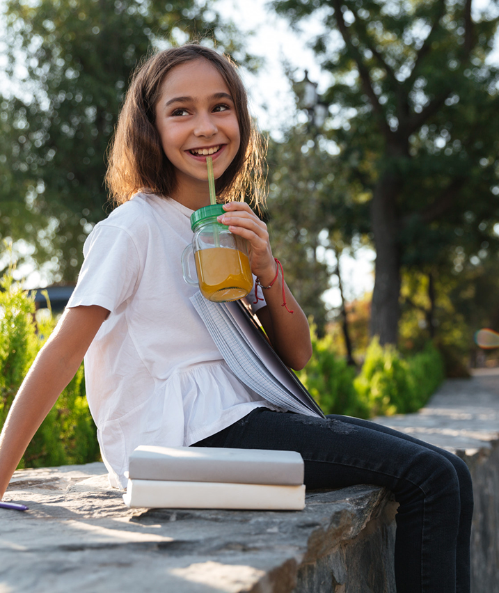 Smiling girl sitting outside with books and drinking juice from a jar, enjoying a sunny day