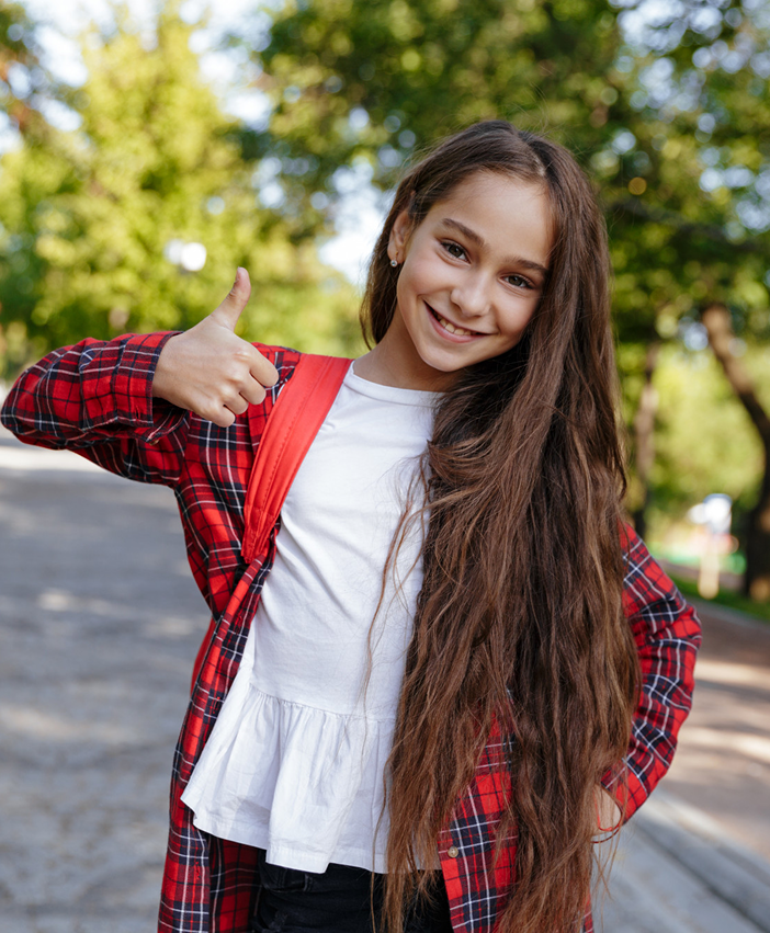 Smiling young girl outdoors giving a thumbs up while wearing a red plaid shirt and backpack