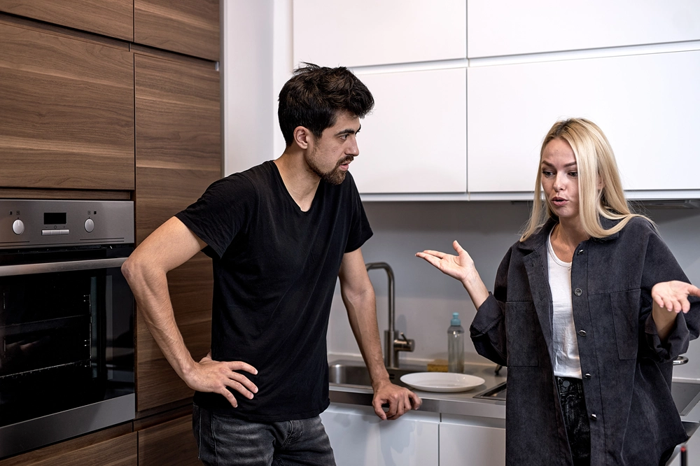Couple standing in kitchen having an argument, with the man looking frustrated and the woman gesturing defensively