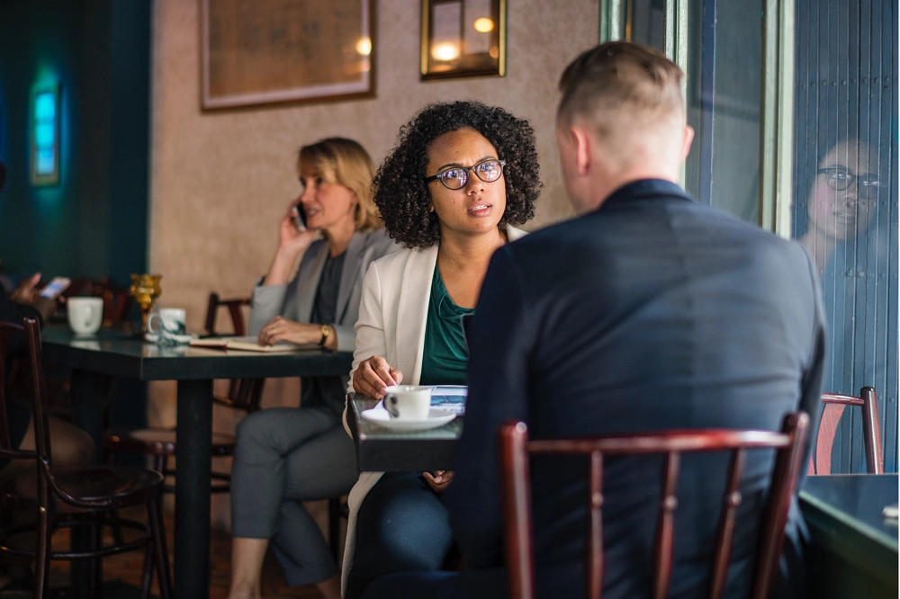 Two people sitting at a café table having a serious conversation, with coffee cups in front of them and others in the background