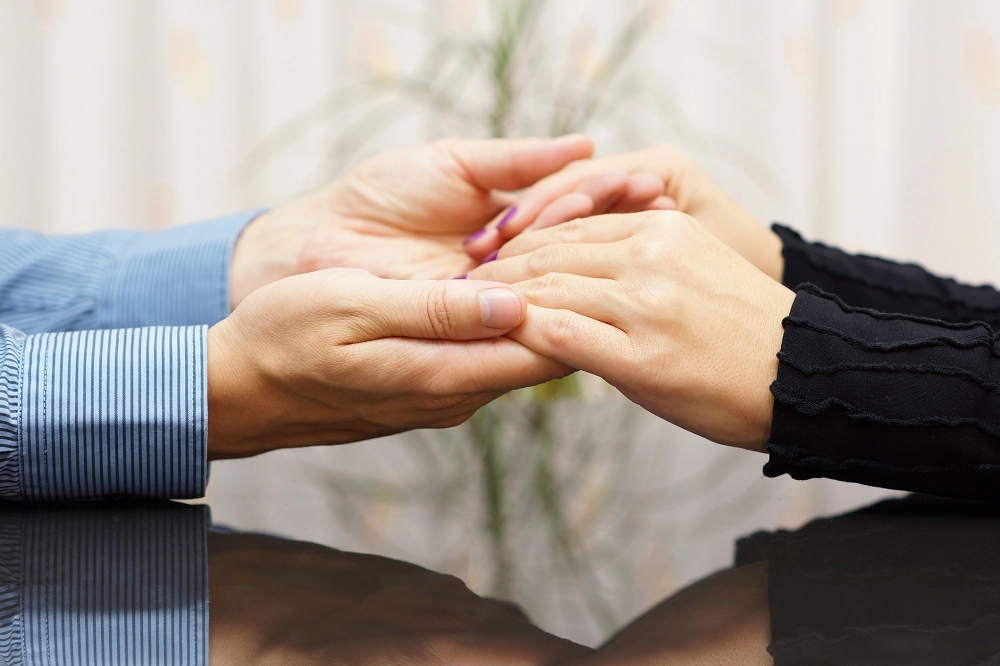 Close-up of two people sitting across from each other, gently holding hands in a comforting and supportive gesture