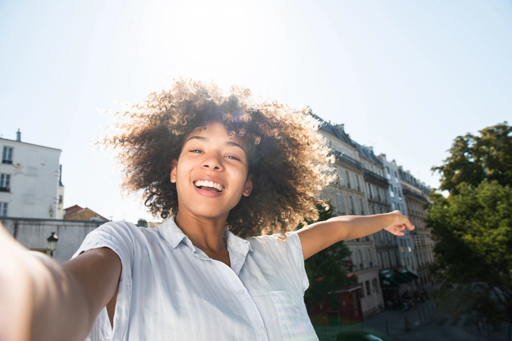 Smiling young woman taking a selfie outdoors on a sunny day with city buildings in the background