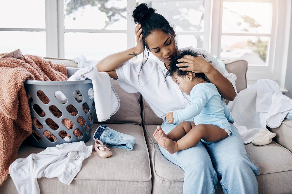 Mother comforting her young child while sitting on a couch surrounded by laundry and household clutter