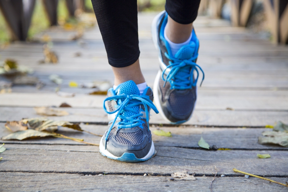 Close-up of a person walking outdoors on a wooden path wearing blue running shoes and black leggings, with fallen leaves scattered on the ground
