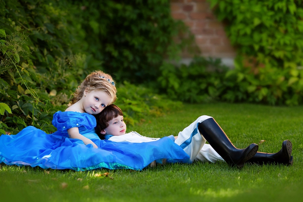 Two children dressed as a princess and prince resting on the grass in a garden