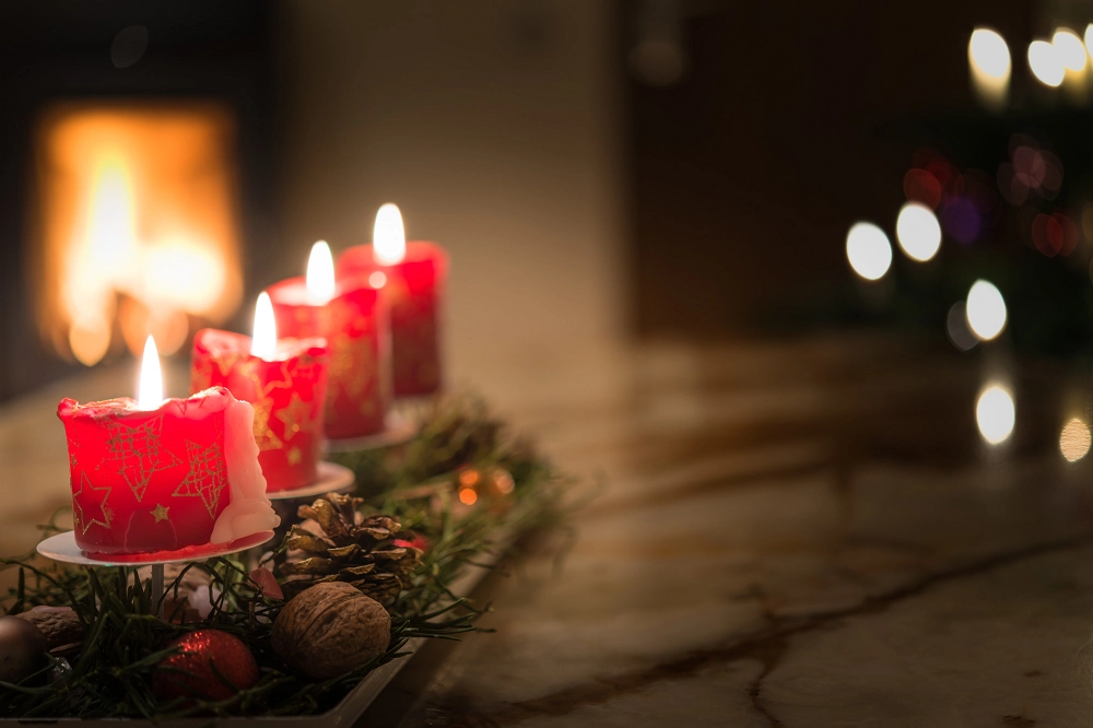 Four red Christmas candles burning on a festive wreath with pinecones and greenery beside a warm fireplace