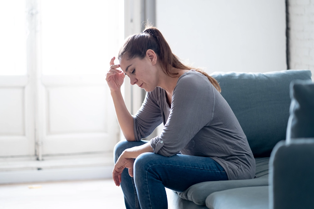 Woman sitting on couch with head down and hand on forehead, appearing sad or stressed