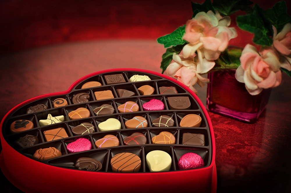 Heart-shaped box filled with assorted chocolates next to a vase of pink roses on a red surface