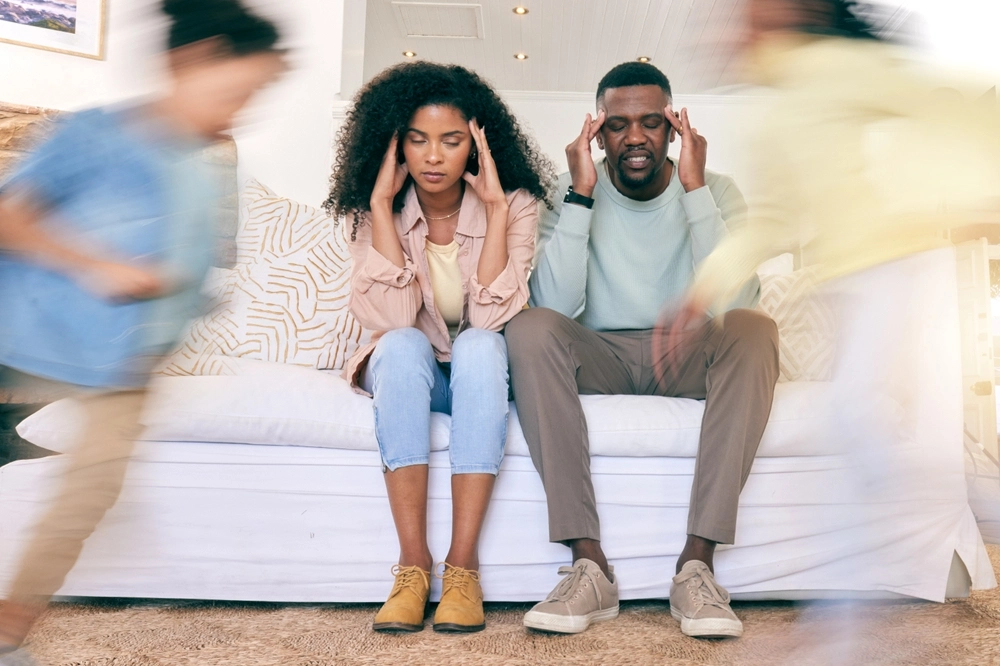 Stressed parents sitting on a couch holding their heads while children run around the room