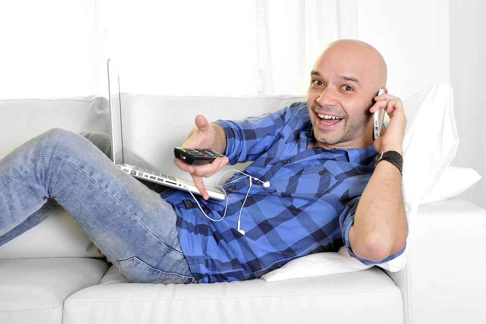 Smiling man multitasking on a couch with a laptop, phone, and TV remote in hand