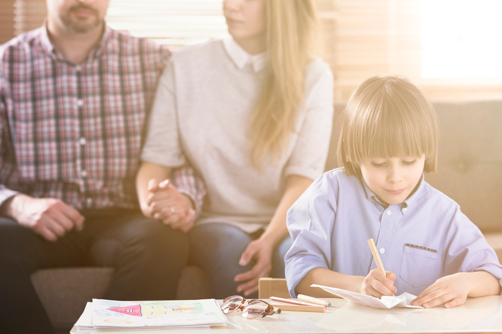 Young boy drawing at a table while his parents sit together in the background holding hands