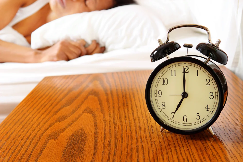 Black alarm clock on a wooden nightstand showing 7 o’clock with a person resting in bed