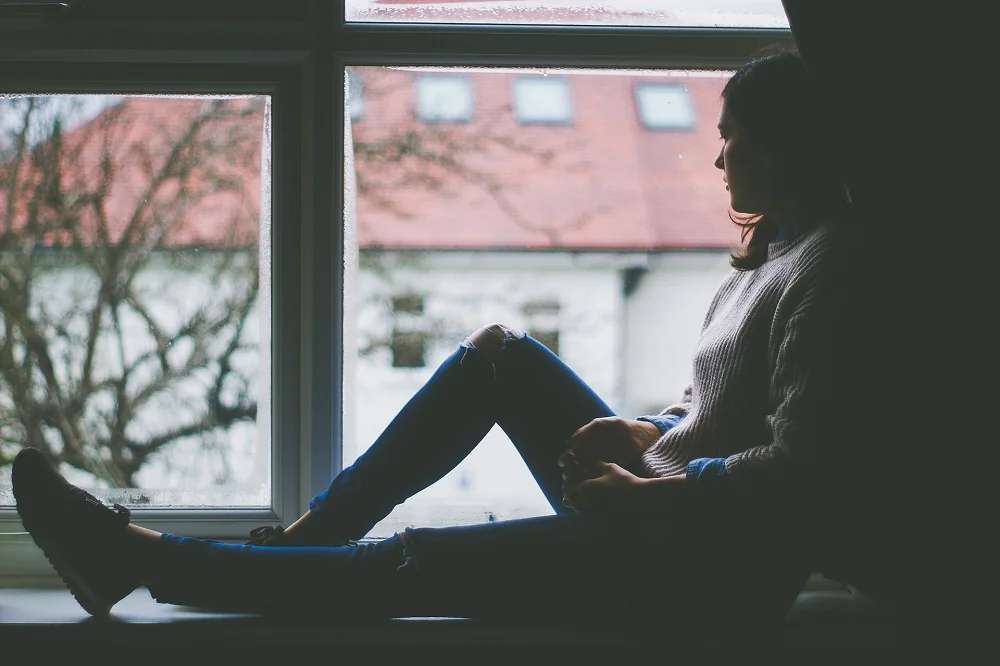 Person sitting by a window looking outside thoughtfully on a cloudy day, conveying a reflective mood