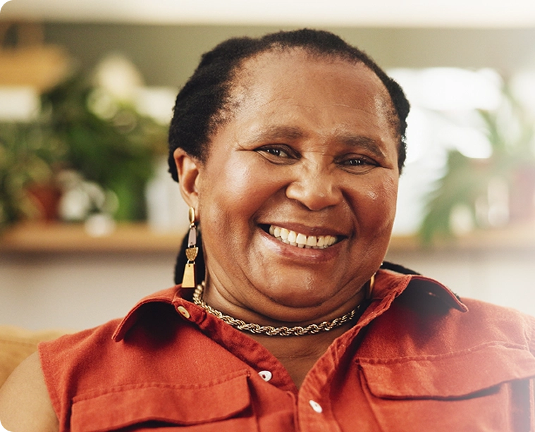 Smiling woman sitting indoors wearing an orange shirt and gold jewelry, radiating warmth and happiness