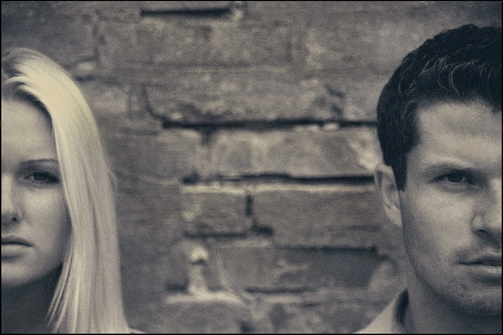 Close-up black-and-white photo showing a man and a woman standing apart with serious expressions against a brick wall