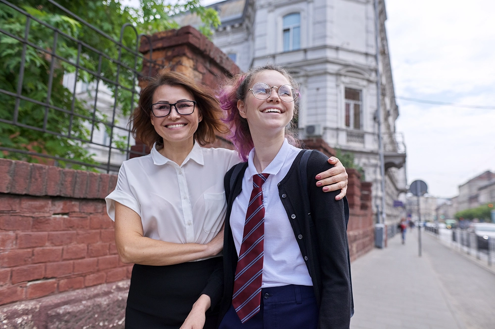 Smiling woman with arm around teenage girl in school uniform standing on a city sidewalk