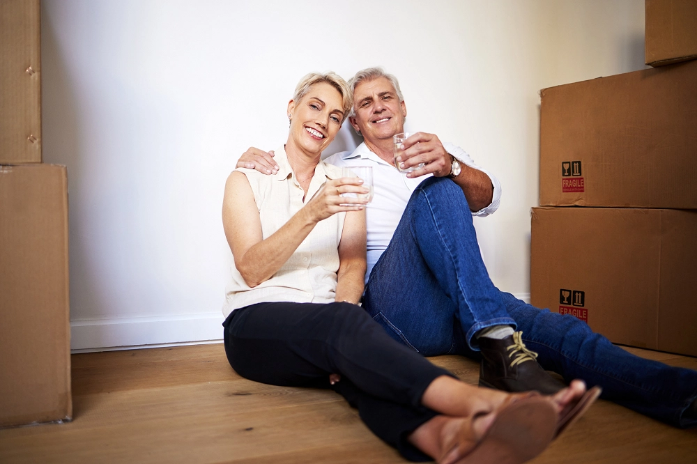 Smiling older couple sitting on the floor surrounded by moving boxes, holding drinks and relaxing together