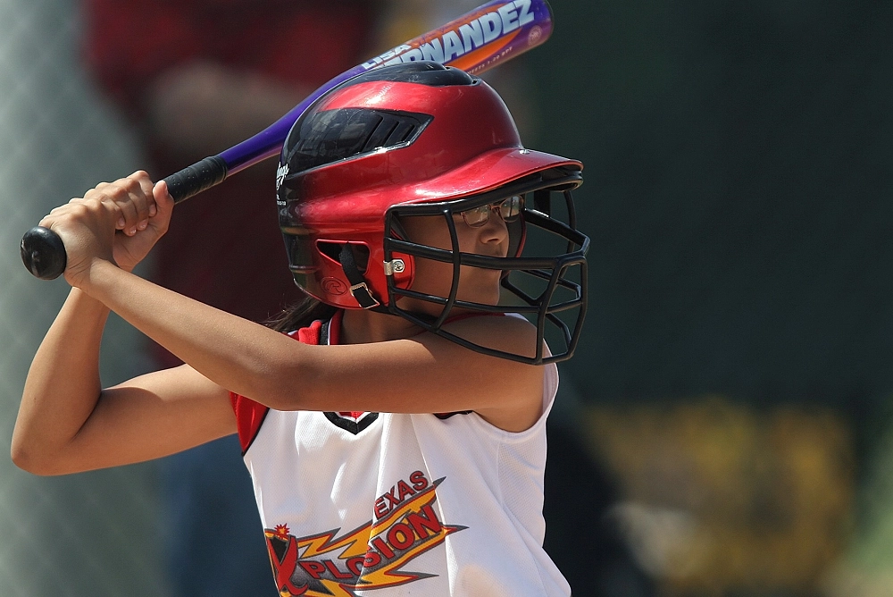 Girl in a red helmet and white jersey holding a bat, preparing to swing during a softball game