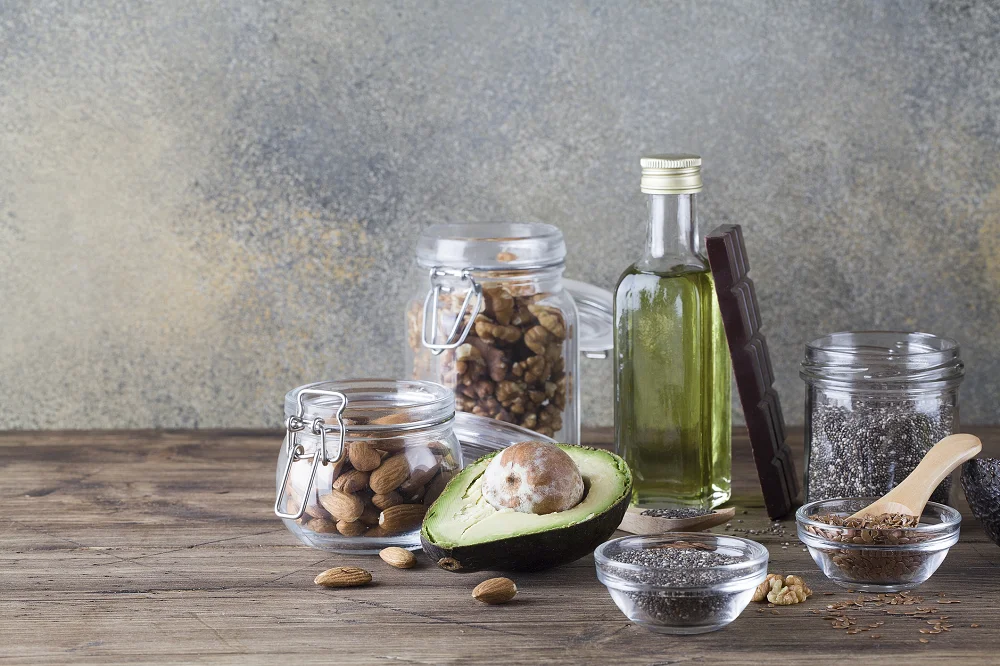 Healthy fats including avocado, nuts, seeds, olive oil, and dark chocolate arranged on a wooden table