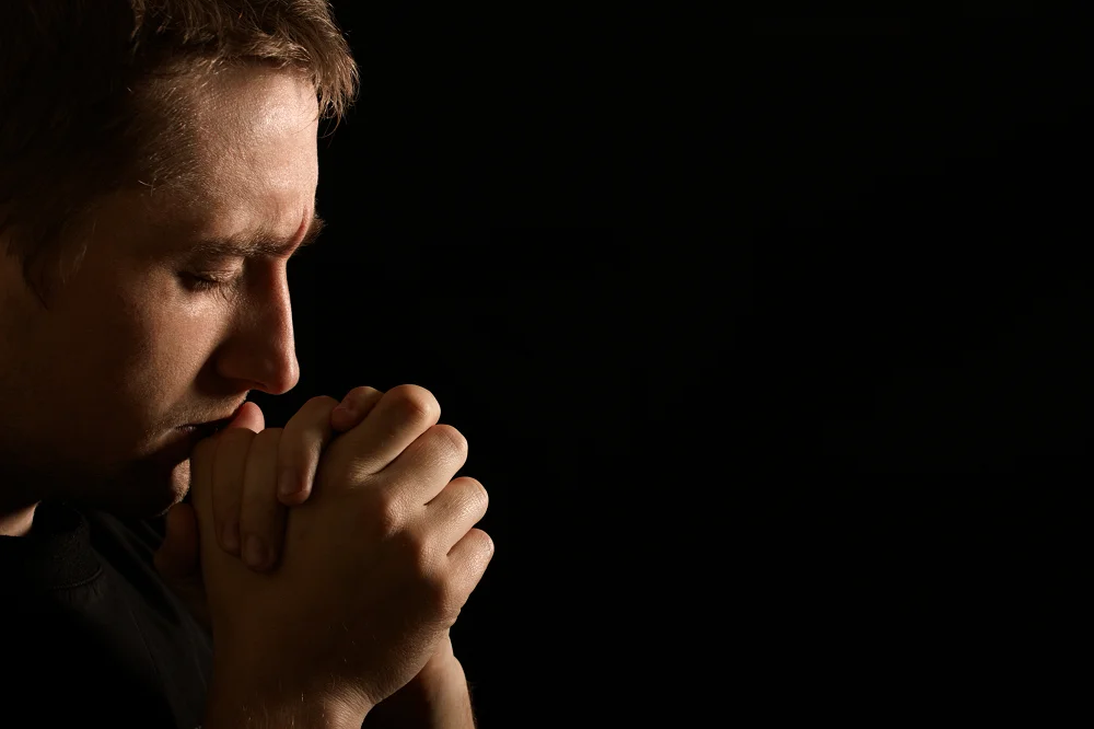 Man with closed eyes holding his hands together in deep thought or prayer against a dark background