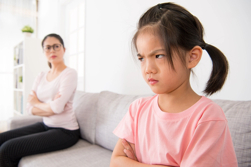 Young girl looking upset while sitting on a couch with her mother in the background crossing her arms