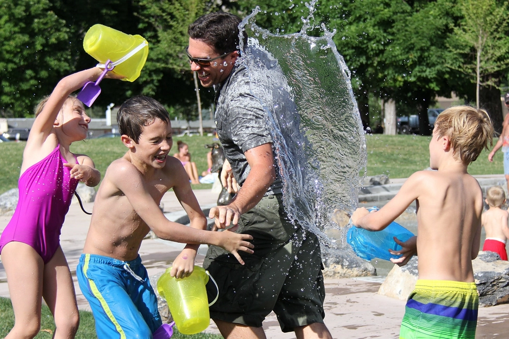 Children and an adult laughing and splashing each other with water buckets at a sunny outdoor park