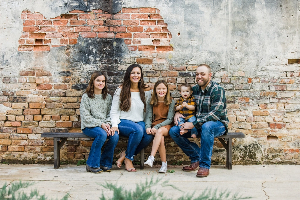 Smiling family of five sitting on a bench in front of a rustic brick wall, posing together for a photo
