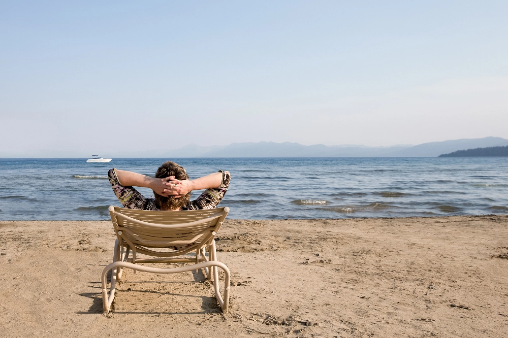 Person relaxing in a chair on a sandy beach, looking out at the calm ocean and distant mountains