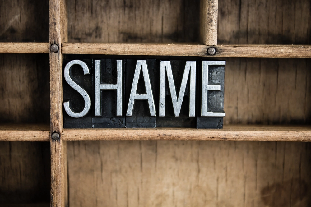 The word “SHAME” spelled out in metal block letters arranged on a wooden shelf