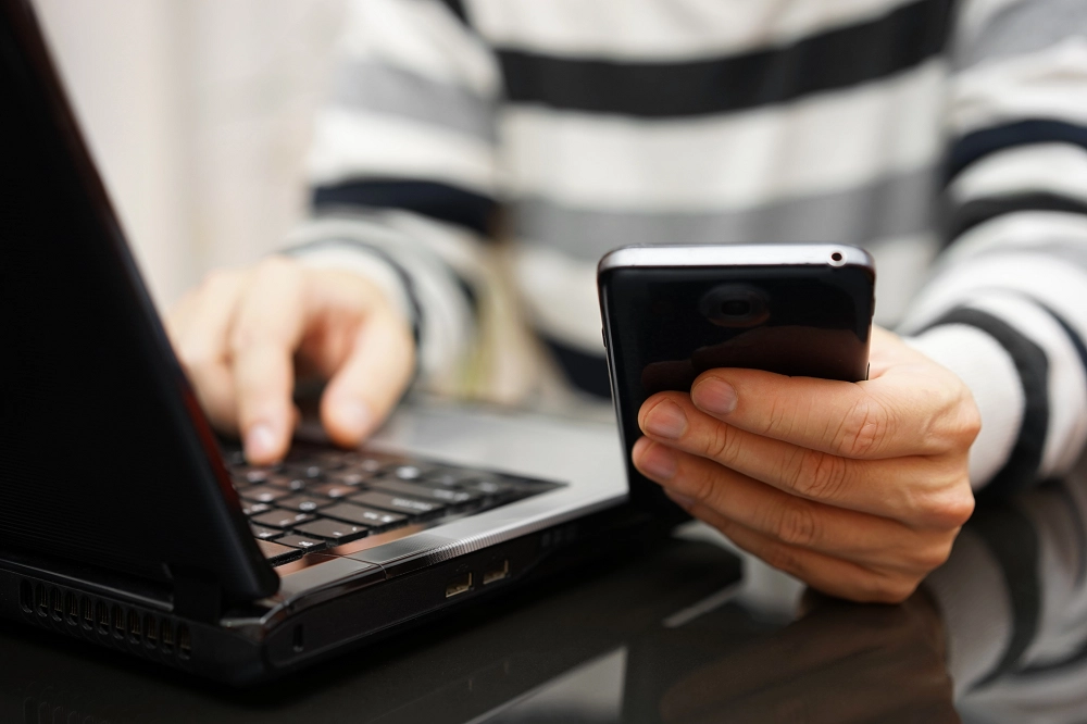 Person using smartphone and laptop simultaneously, representing multitasking and online communication