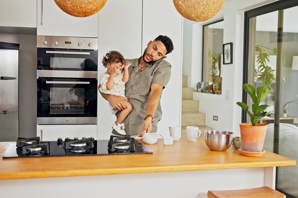 Father holding his young daughter while preparing food in a modern kitchen, showing a busy parenting moment