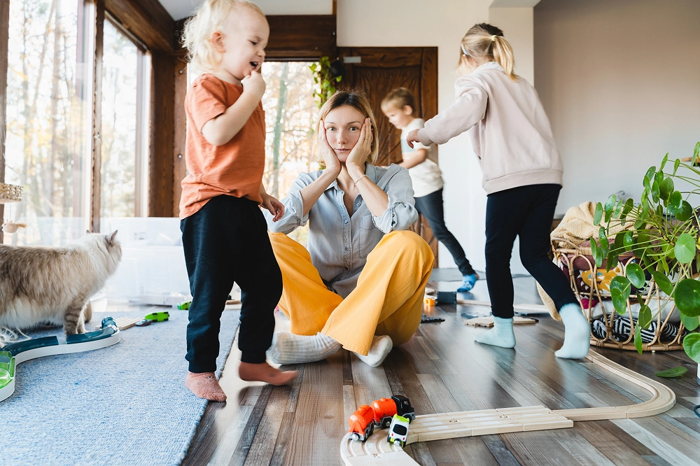 Overwhelmed mother sitting on the floor with children playing around her in a messy living room