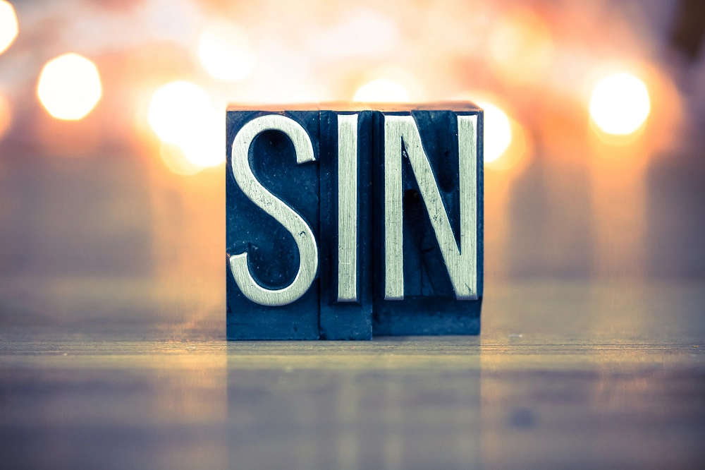 The word “SIN” in bold metal block letters on a wooden surface with blurred lights in the background