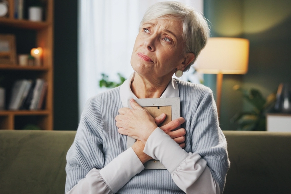 Elderly woman sitting on a couch holding a framed photo to her chest, looking thoughtful and emotional