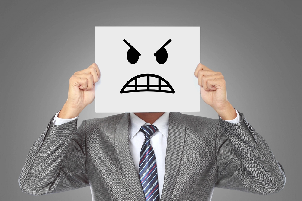 Person in suit holding paper with angry face drawing in front of their head, symbolizing hidden anger or frustration