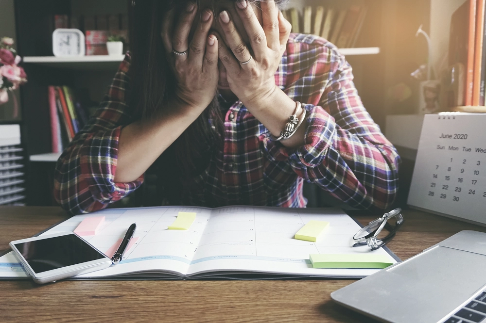 Person sitting at a desk with head in hands, surrounded by a planner, phone, and laptop, appearing stressed