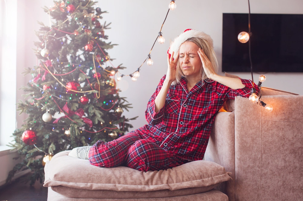 Woman in red pajamas and Santa hat holding her head with stress near a decorated Christmas tree