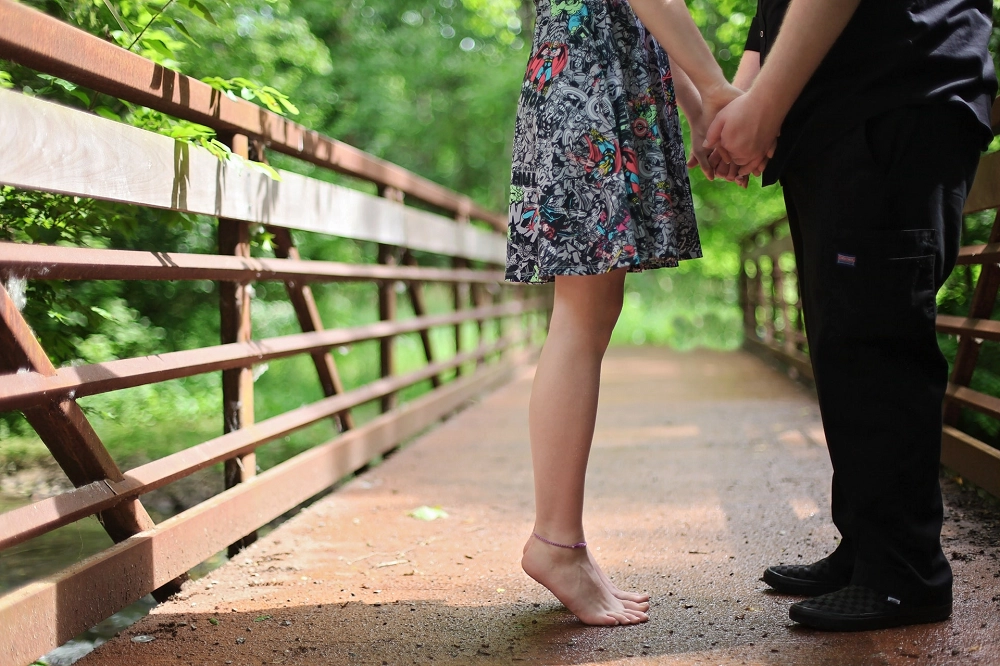 Couple holding hands while standing close together on a wooden bridge surrounded by greenery