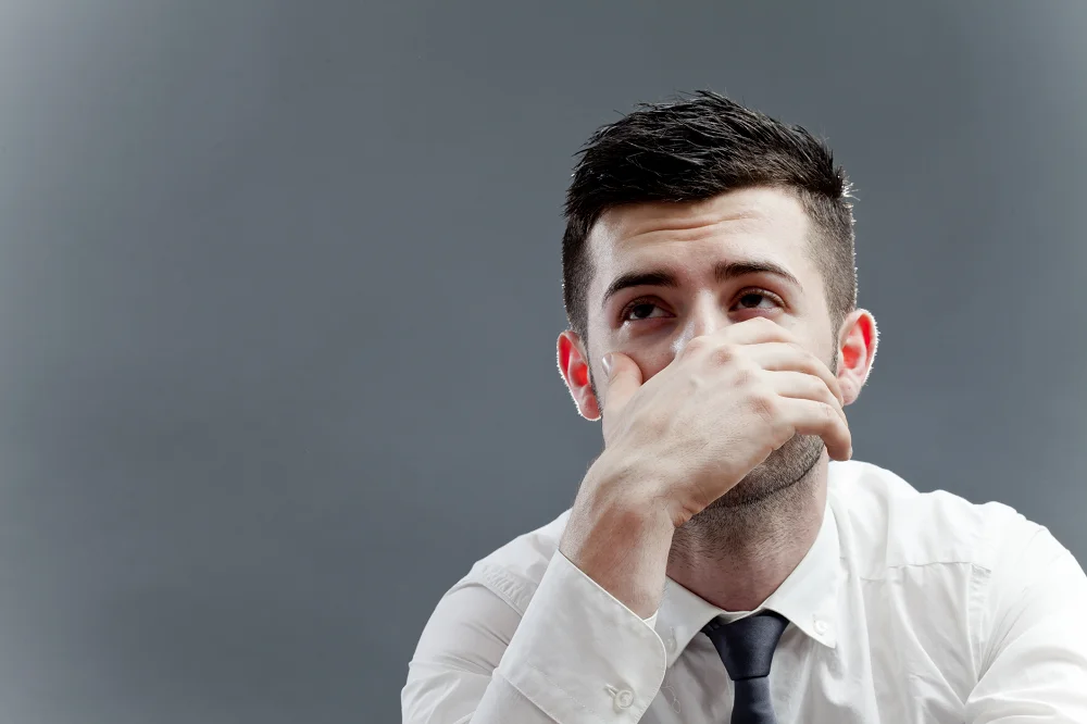 Man in a white shirt looking thoughtful with his hand covering part of his face against a gray background