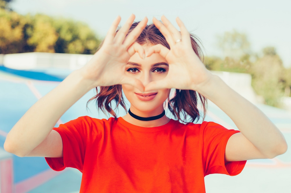 Smiling young woman in a red shirt forming a heart shape with her hands outdoors