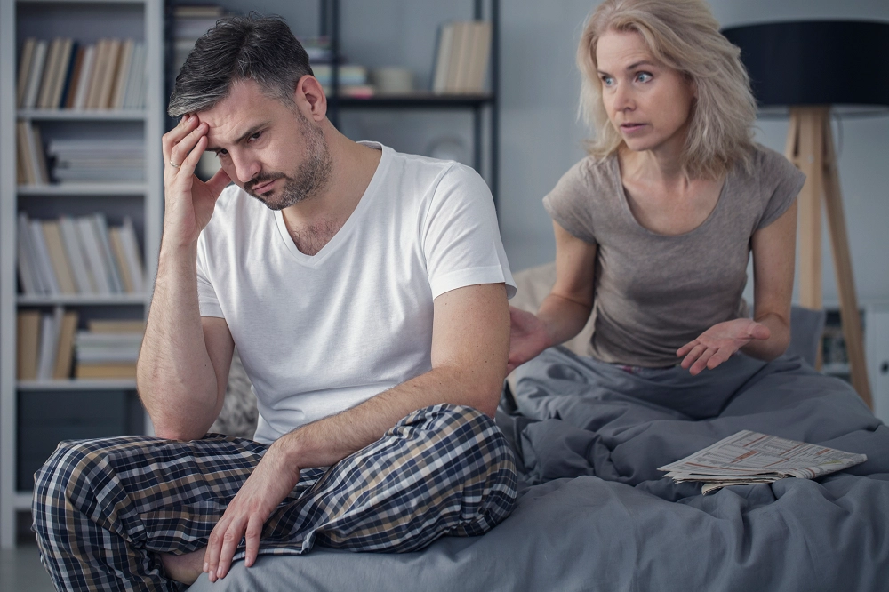 Couple sitting on a bed having a tense conversation, with the man looking stressed and the woman gesturing toward him