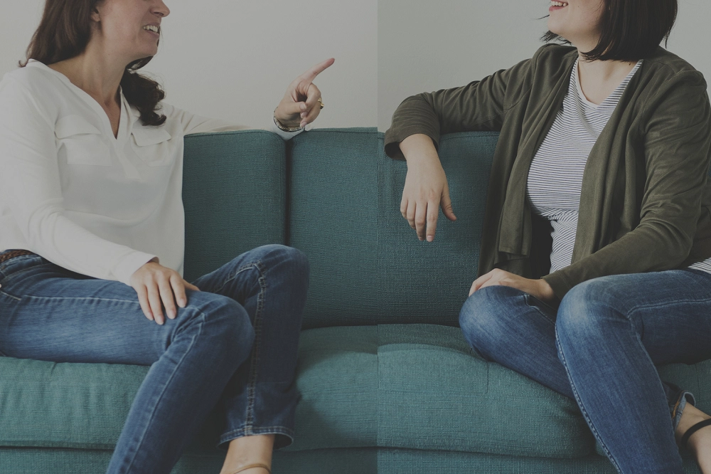 Two women sitting on a couch engaged in conversation, one gesturing while the other listens attentively