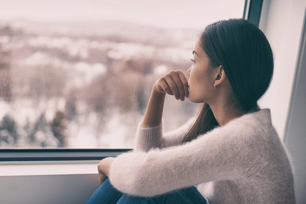 Woman sitting by window looking outside thoughtfully, reflecting or feeling sad on a winter day