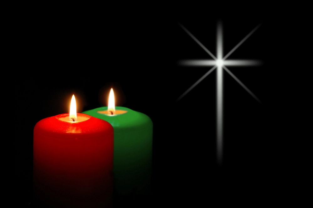 Red and green candles burning beside a glowing white cross on a black background, symbolizing faith and remembrance
