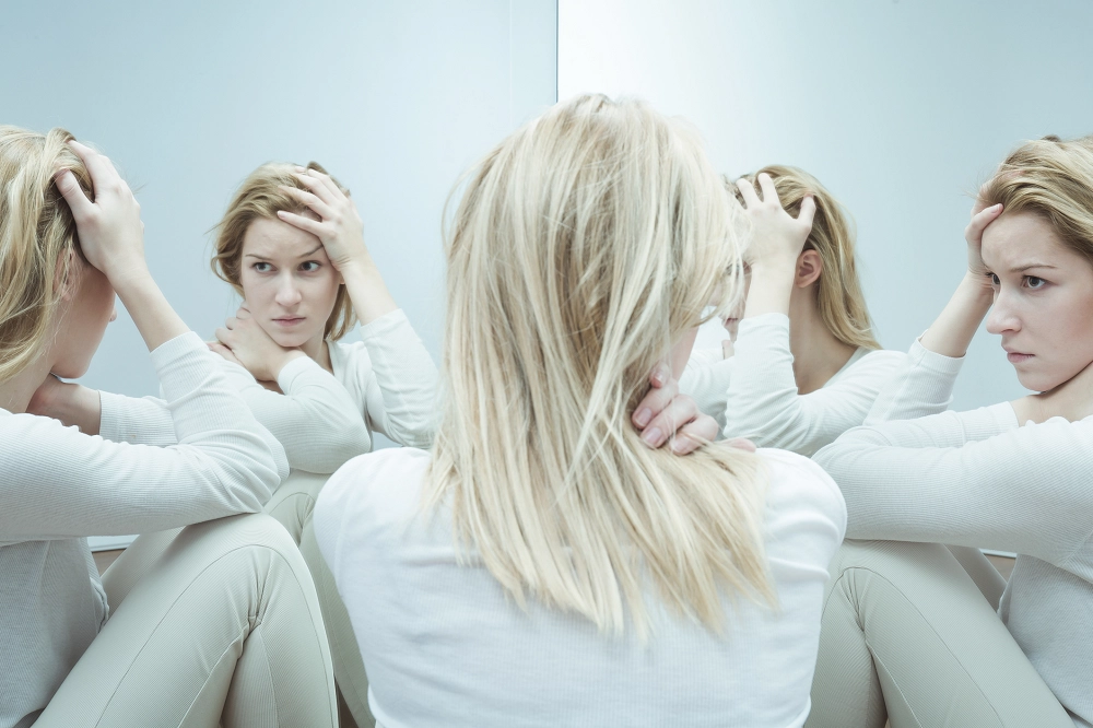 Woman sitting with head in hands surrounded by reflections, symbolizing anxiety, inner conflict, or mental distress