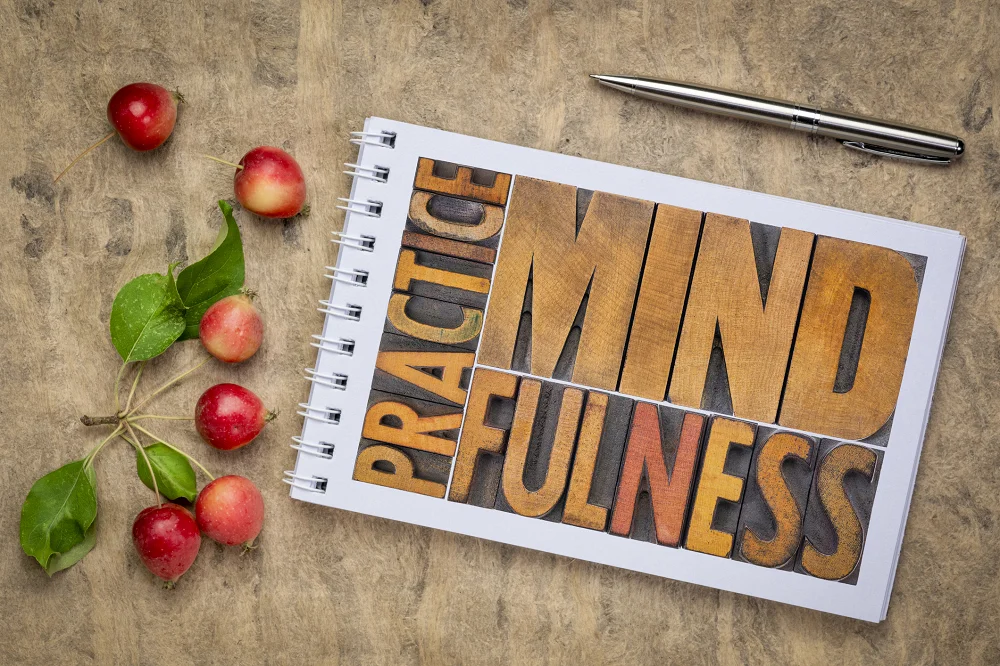 Notebook with the words ‘Practice Mindfulness’ in wooden block letters beside small red apples and a pen on a rustic surface