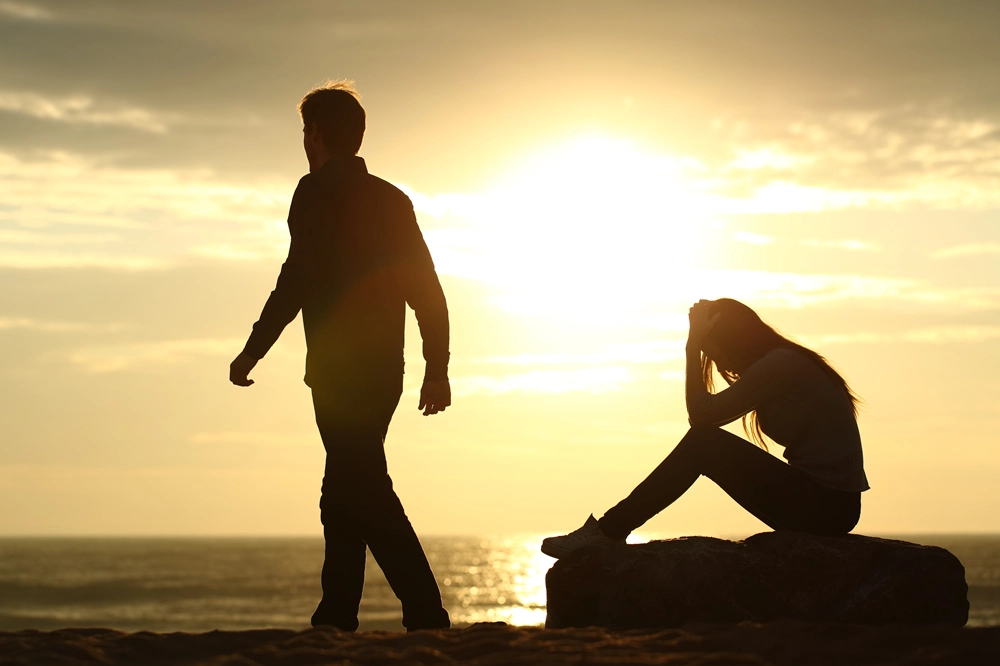 Silhouette of man walking away from seated woman holding her head in distress at sunset